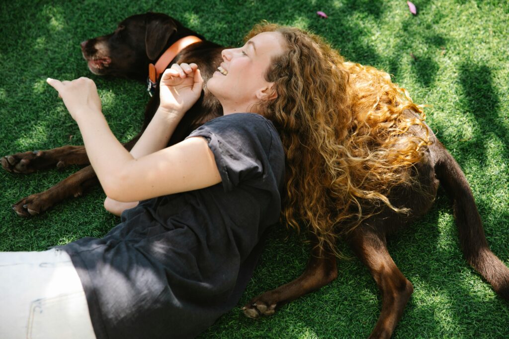 From above side view of glad female with curly hair lying with bird dog on meadow in sunlight