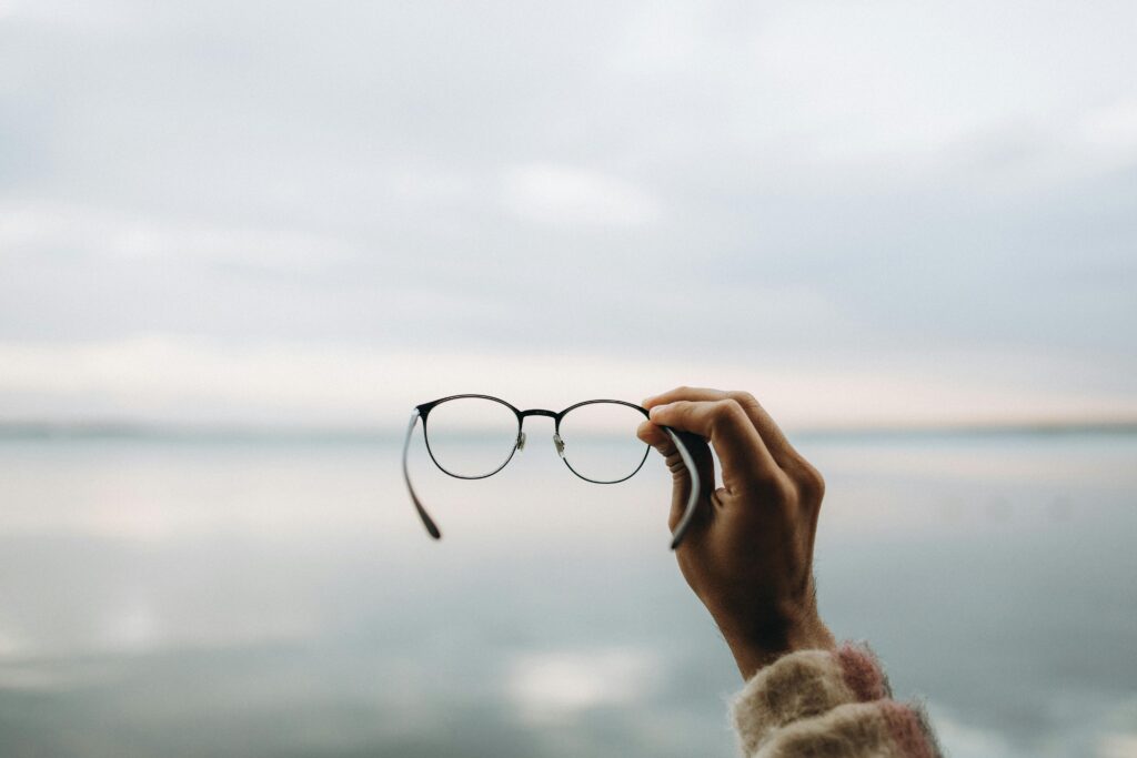 A hand holding eyeglasses with a calm lake and soft sky in the background, evoking tranquility.